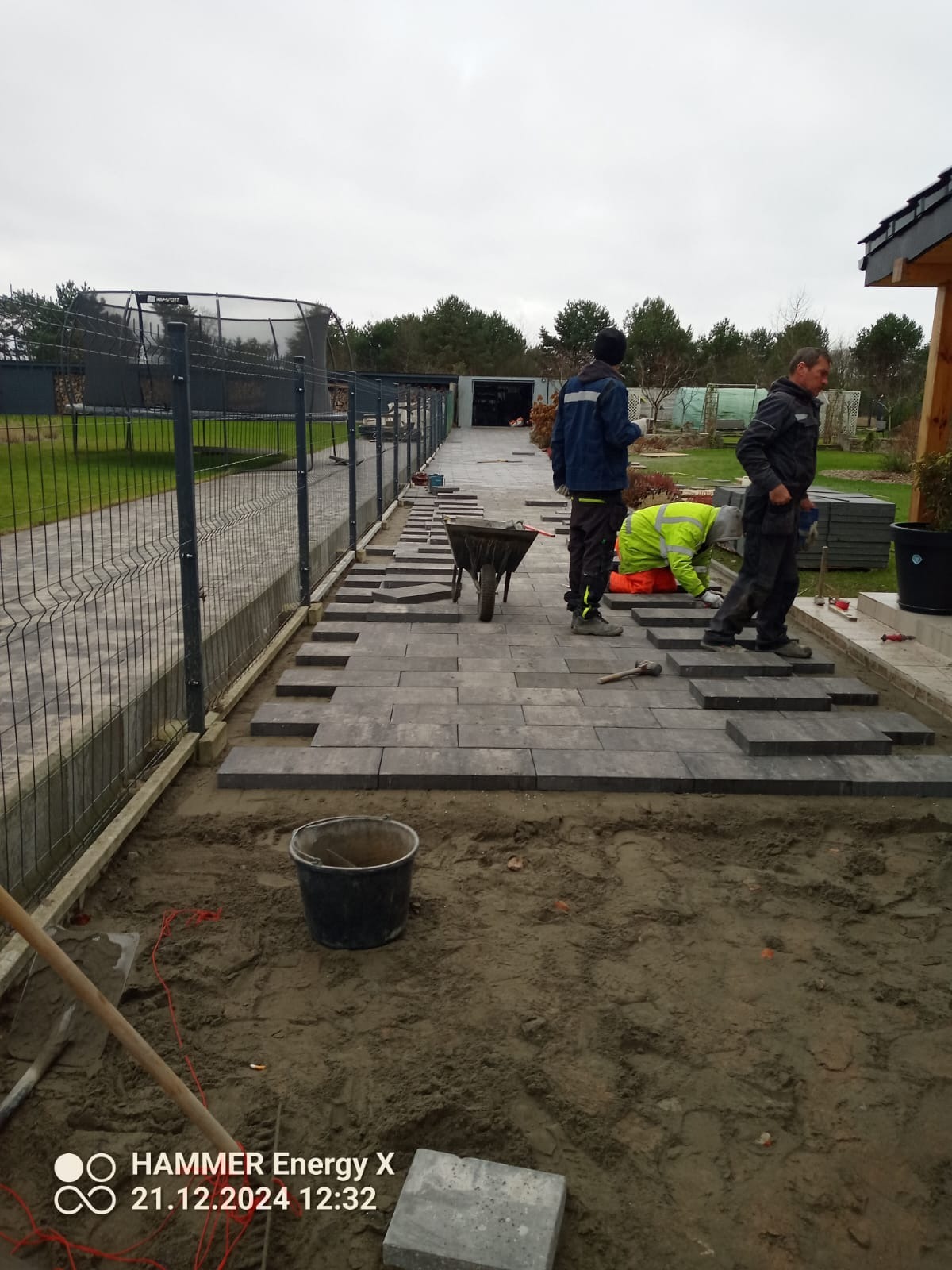Trzech pracowników układa szarą kostkę brukową na posesji, widoczny fragment ogrodzenia, taczka i narzędzia. Częściowo ułożony chodnik prowadzi w głąb działki.
