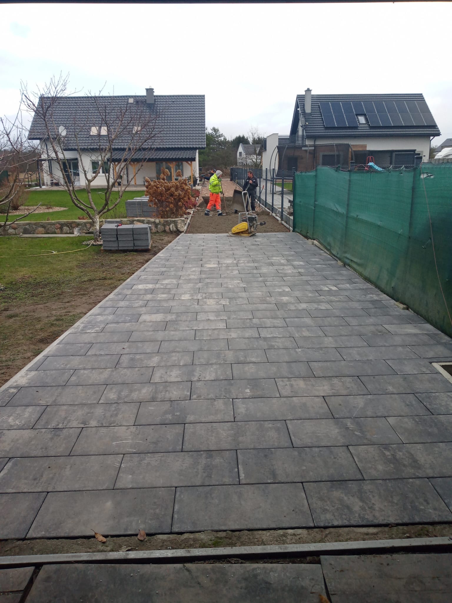 Układanie szarej kostki brukowej na posesji. Widoczni dwaj pracownicy, jeden z zagęszczarką. W tle domy jednorodzinne i zielona siatka ogrodzeniowa.