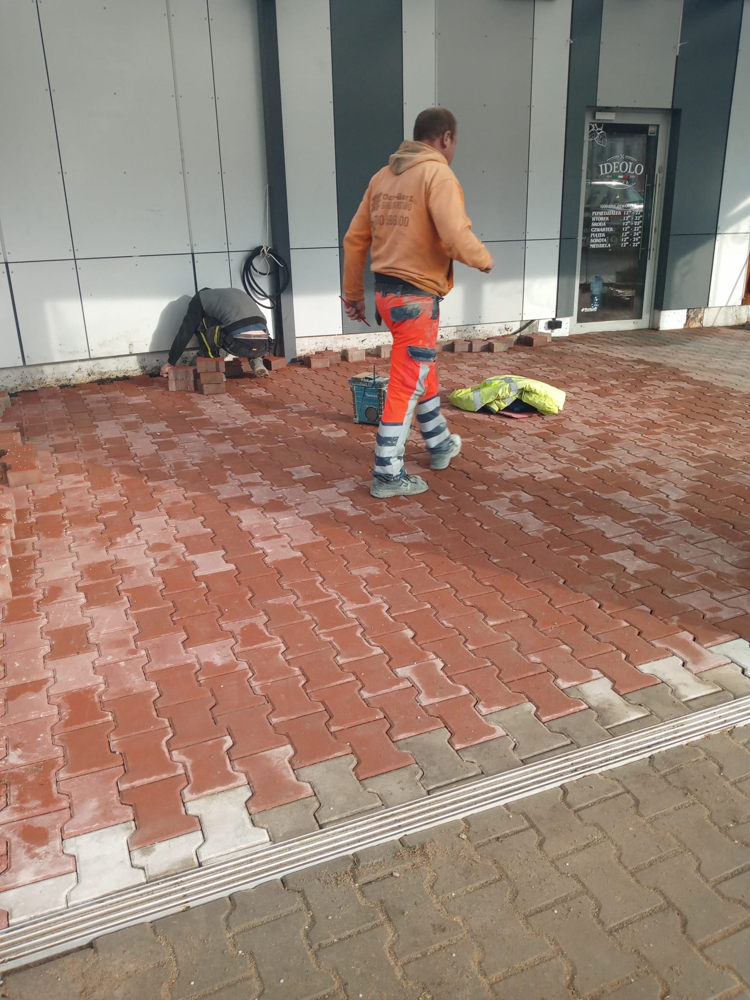 Układanie czerwonej kostki brukowej typu I na zewnątrz budynku. Dwóch pracowników w odblaskowych ubraniach podczas pracy. Częściowo ułożona nawierzchnia.