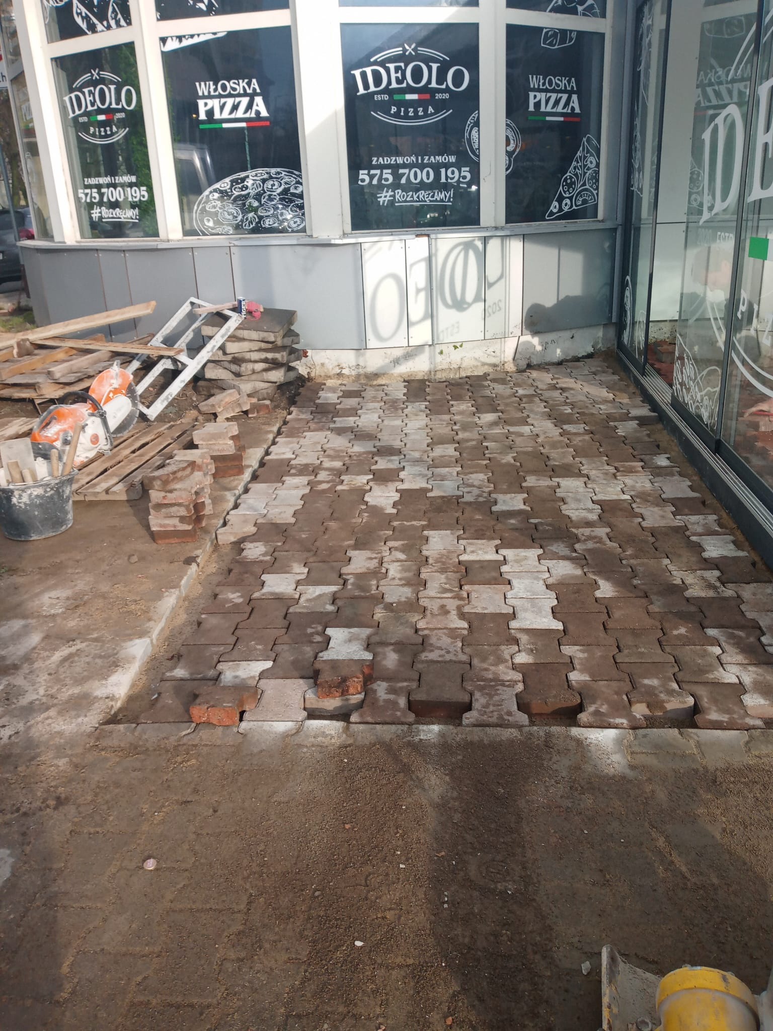 Układanie kostki brukowej typu I przed lokalem gastronomicznym. Widoczne narzędzia i materiały budowlane. Częściowo ułożona nawierzchnia z kostki w odcieniach szarości i brązu.