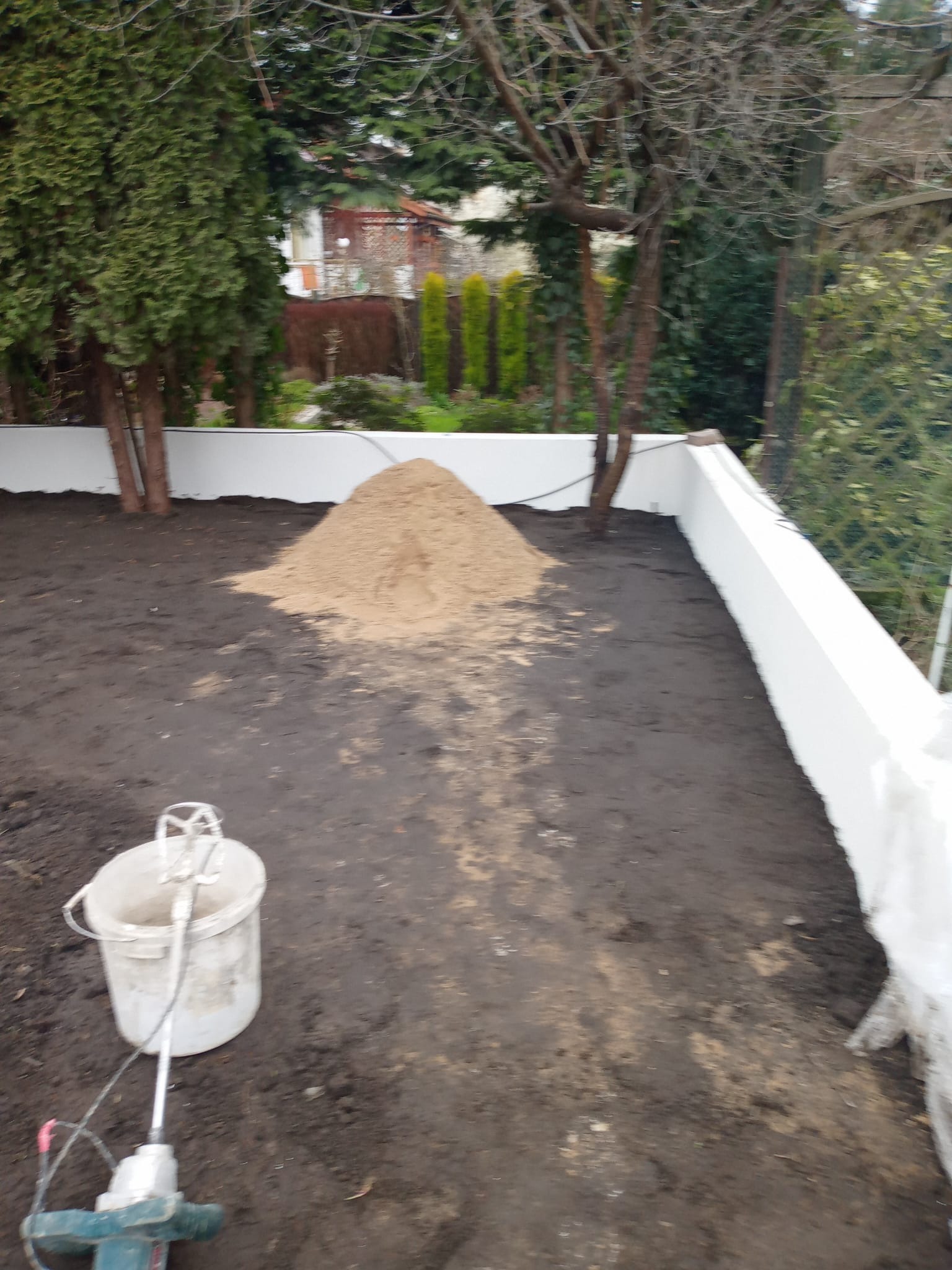 Przygotowanie podłoża: czarna ziemia, piasek i mieszadło budowlane na działce z białym murem. Widoczne drzewa i krzewy w tle. Etap prac ziemnych przed dalszymi pracami.