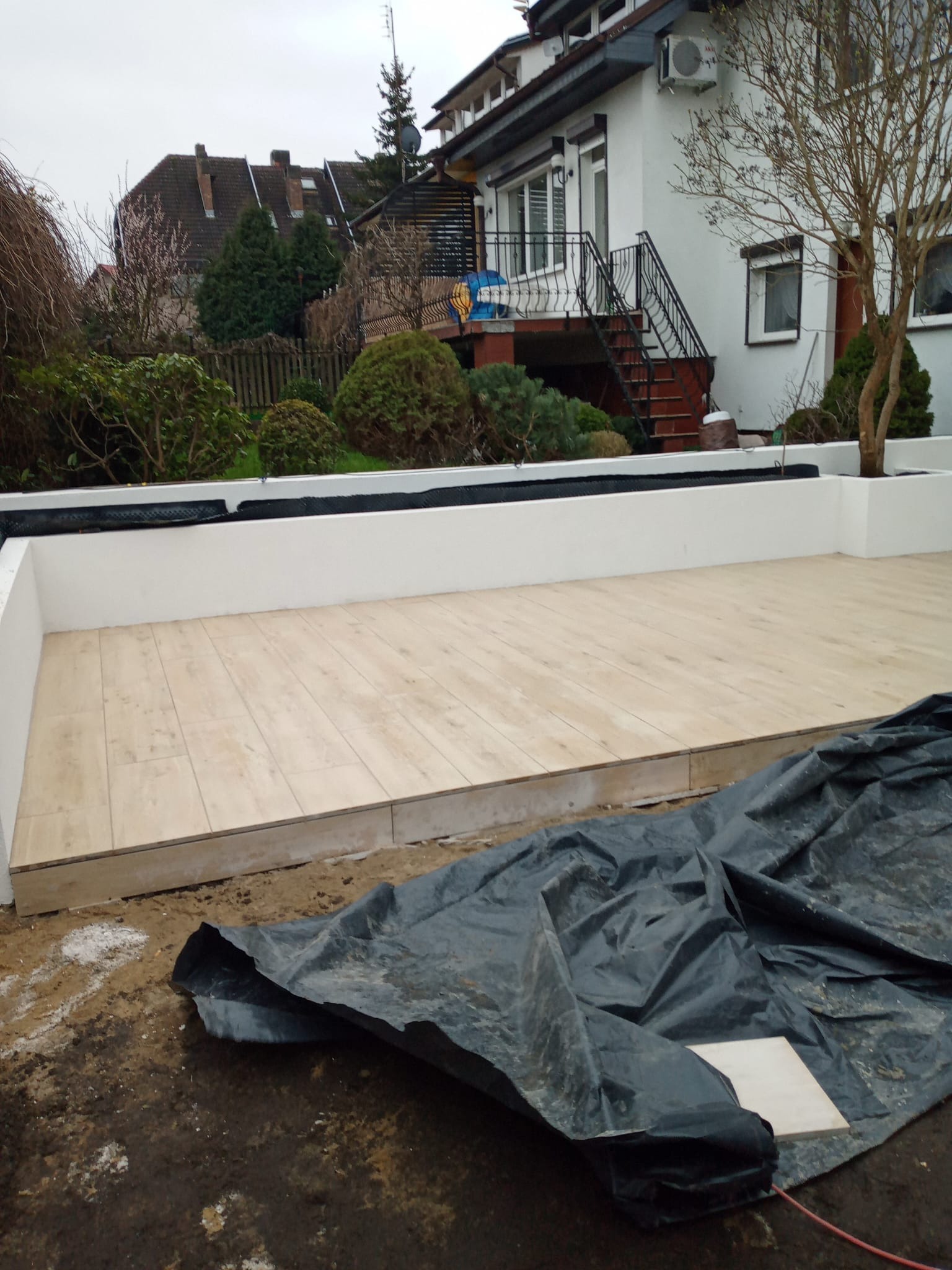 Jasna, drewnopodobna podłoga tarasu w trakcie budowy, otoczona białymi ściankami. W tle dom z czerwonymi schodami i zielenią. Czarna folia budowlana na pierwszym planie.