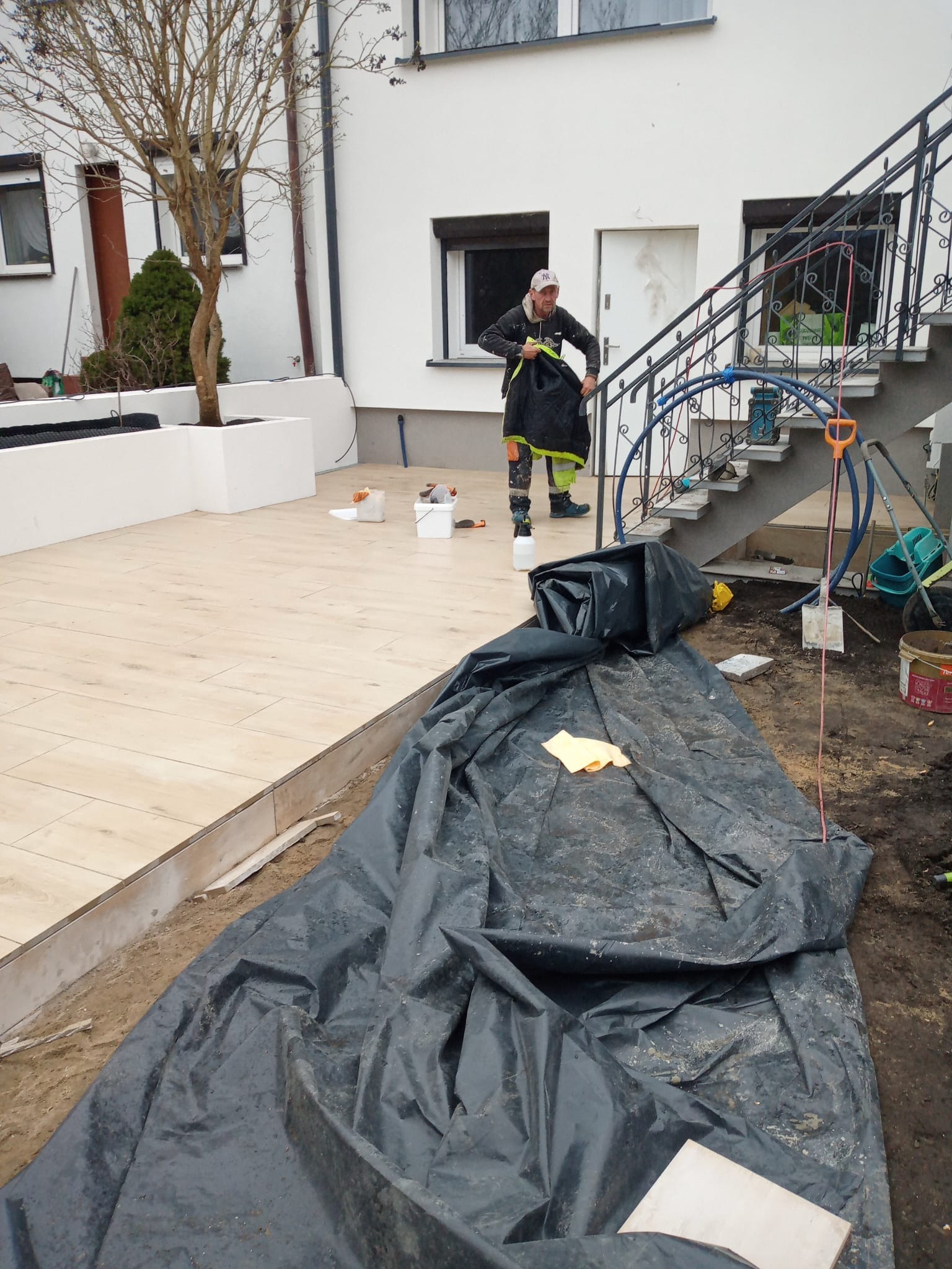 Prace wykończeniowe na tarasie: mężczyzna w stroju roboczym przygotowuje się do dalszych działań. Widoczne płytki imitujące drewno, narzędzia i czarna folia ochronna.