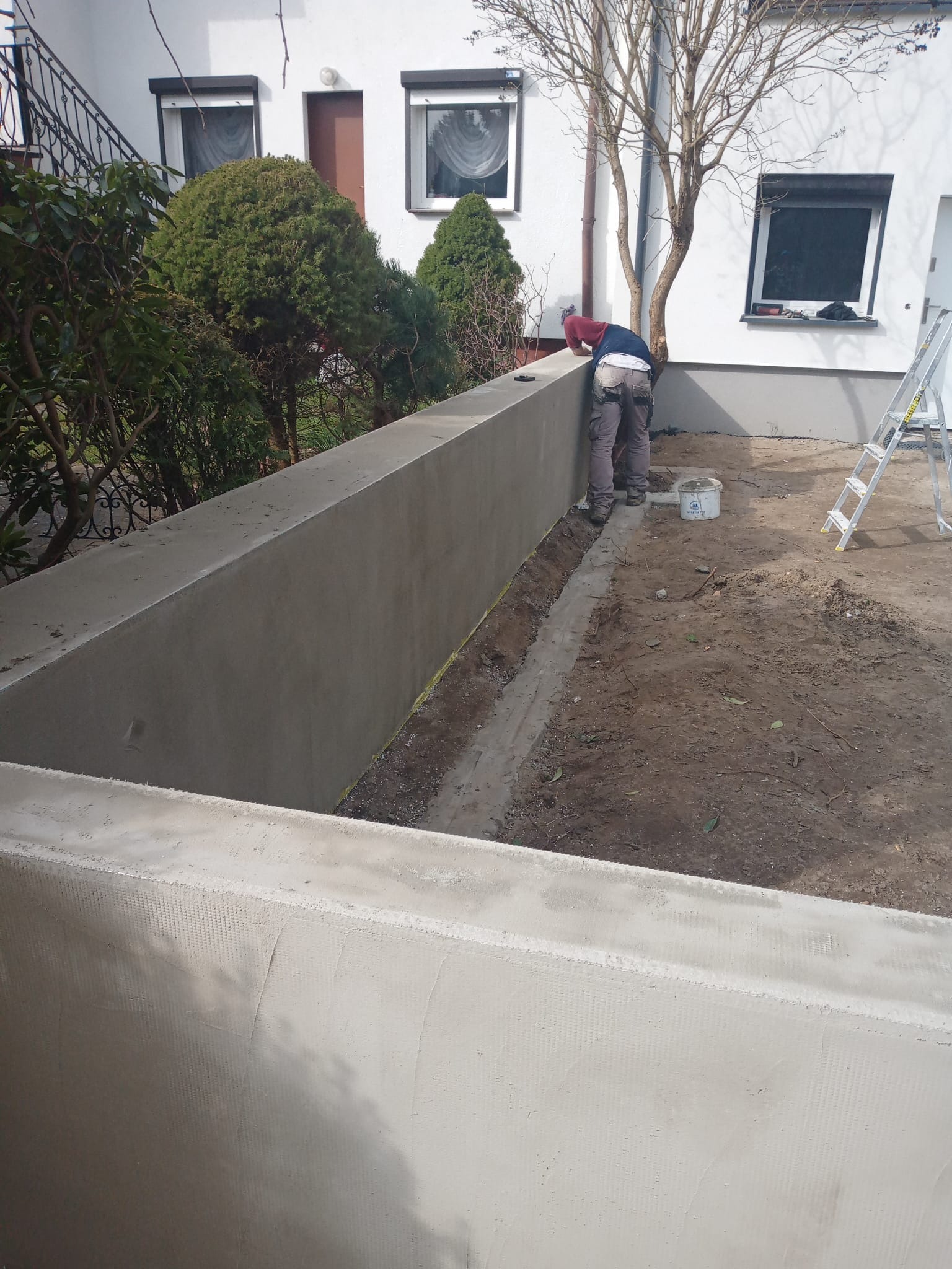 Budowa betonowego murku ogrodowego: pracownik wygładza powierzchnię, widoczne zbrojenie i przygotowany teren pod dalsze prace. Dom w tle.