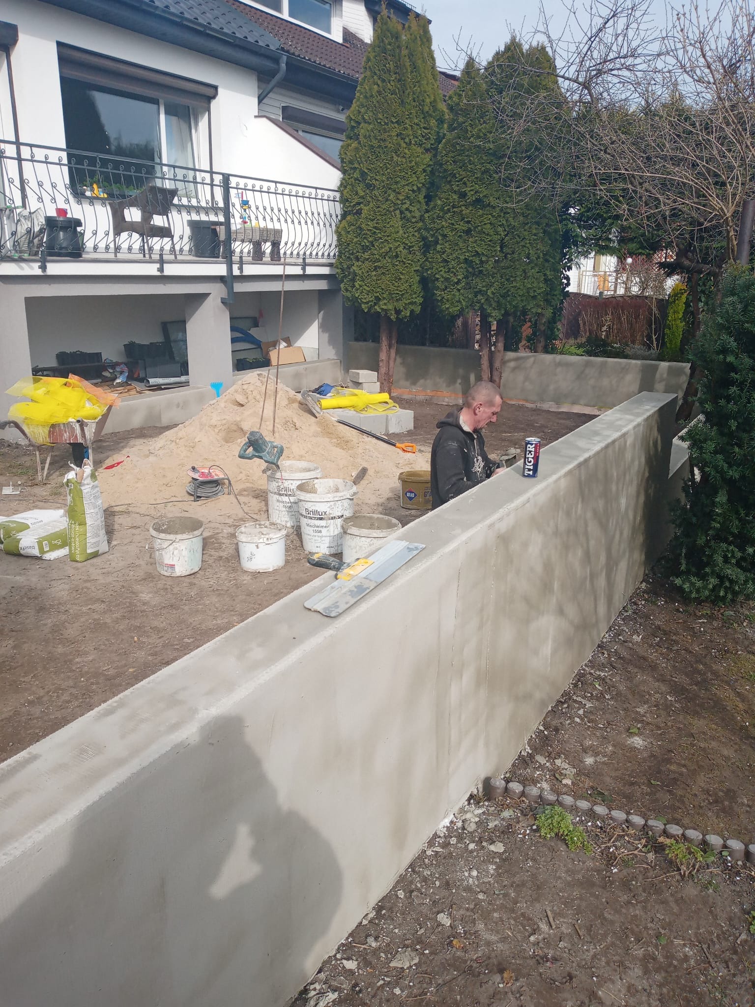 Pracownik wygładza świeżo wylaną, betonową ścianę oporową w ogrodzie. Widoczne narzędzia, worki z zaprawą, piasek i dom w tle. Praca w toku, etap wykończeniowy.
