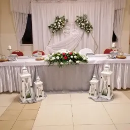 Dekoracja stołu weselnego z białymi lampionami, kompozycją kwiatową z czerwonymi różami i białymi kwiatami, oraz aranżacją z białych materiałów i kwiatów na tle białych zasłon.