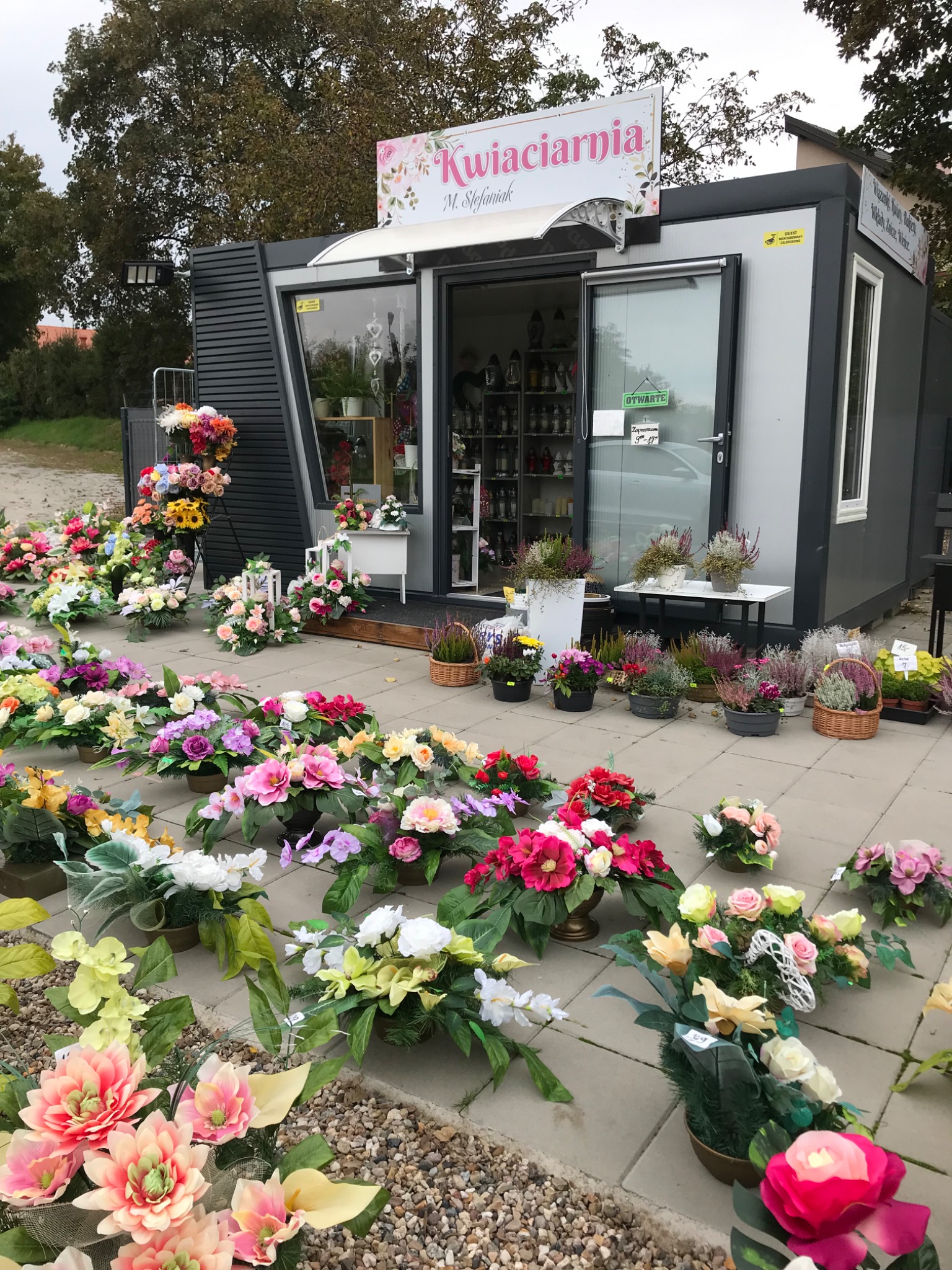 Kwiaciarnia oferująca kompozycje kwiatowe w donicach i koszach, wystawione na zewnątrz przed wejściem do sklepu w kontenerze, z widocznym szyldem 'Kwiaciarnia M. Stefaniak'.