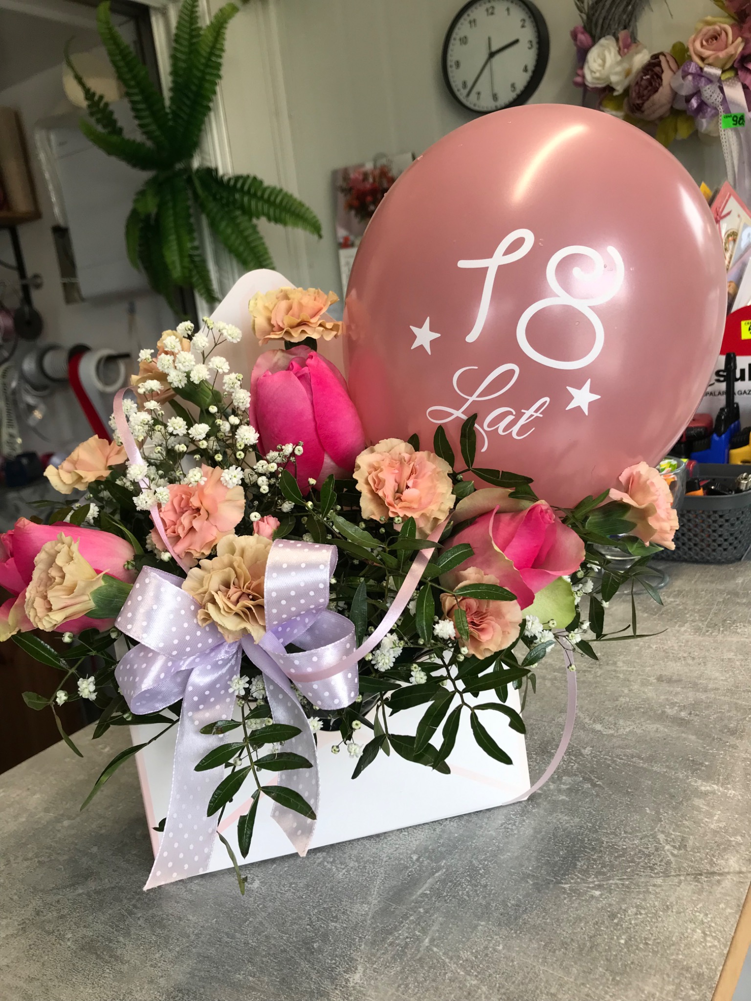 Kompozycja kwiatowa w białym pudełku z różowymi różami, goździkami i gipsówką, ozdobiona fioletową wstążką w białe kropki i różowym balonem z napisem '18 Lat'.