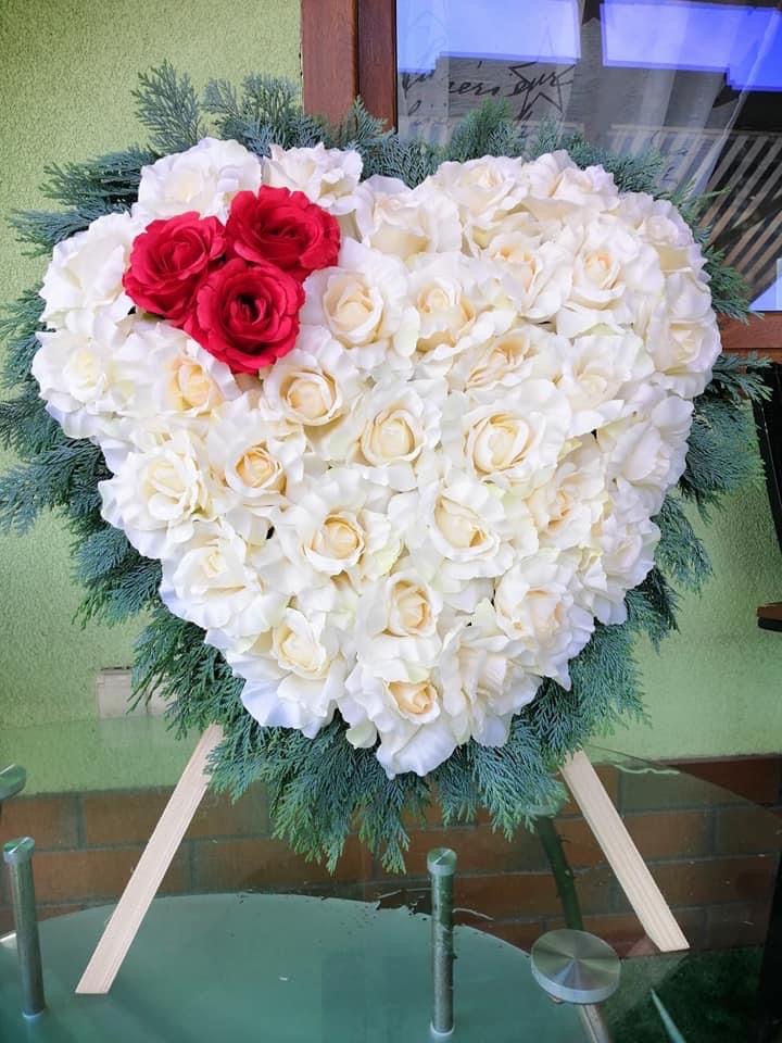 Kompozycja kwiatowa w kształcie serca z białych róż i trzema czerwonymi różami w lewym górnym rogu, otoczona gałązkami igliwia, umieszczona na drewnianym stojaku na szklanym stole.