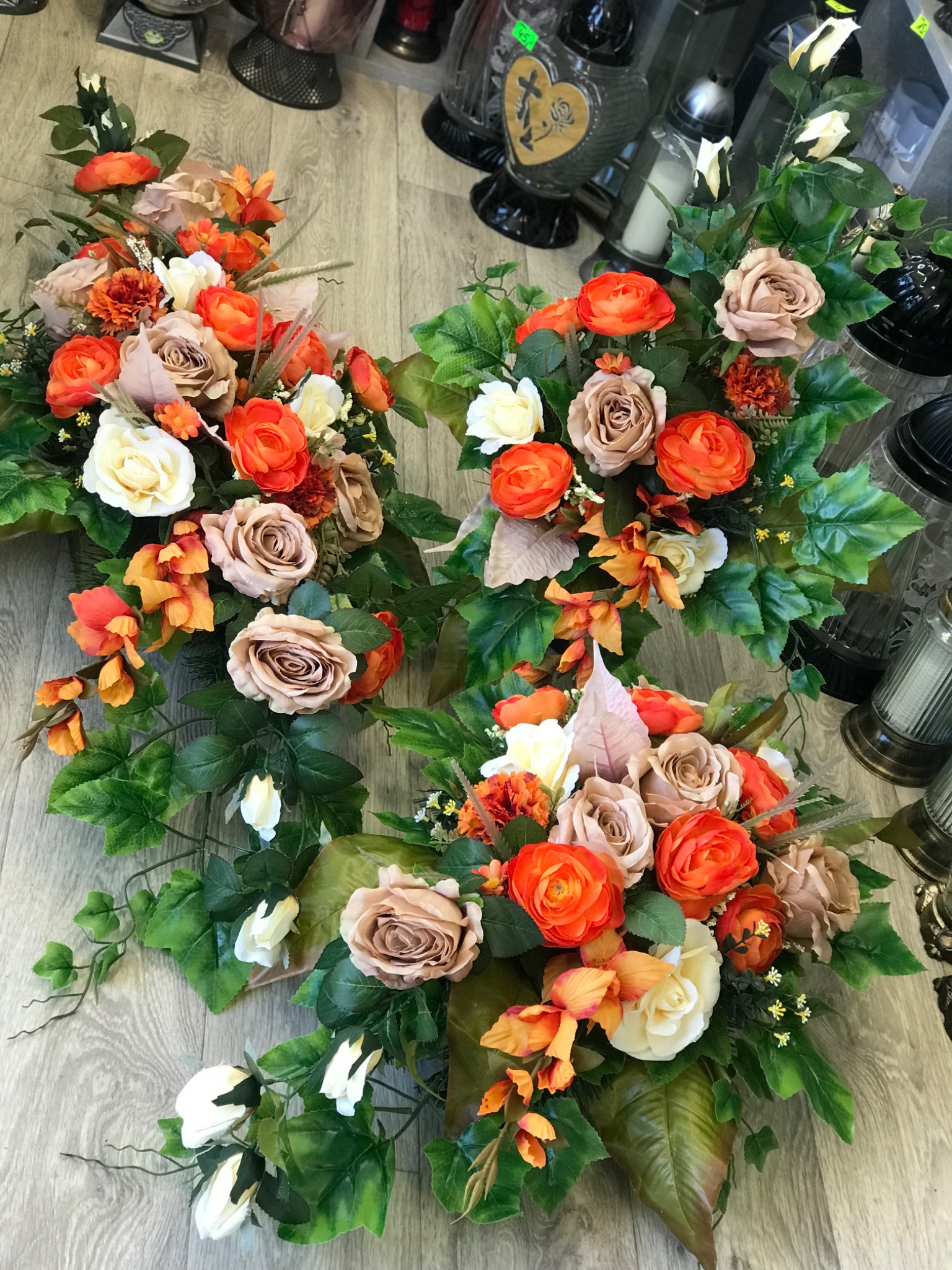 Trzy kompozycje kwiatowe z pomarańczowych, beżowych i białych róż oraz zielonych liści na drewnopodobnym tle, w tle znicze nagrobne.