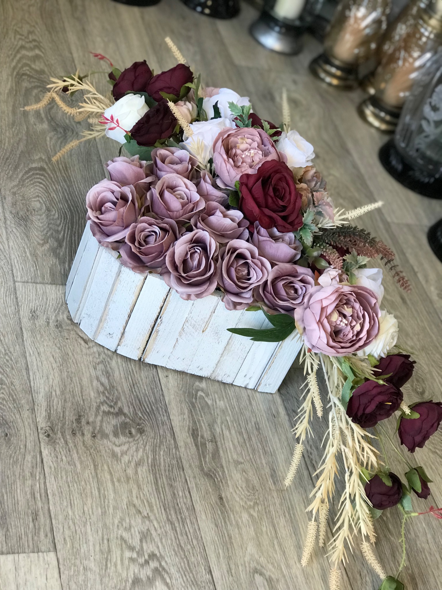 Kompozycja kwiatowa ze sztucznych róż w odcieniach fioletu, bordo i bieli, umieszczona w białej, drewnianej skrzynce na tle drewnianej podłogi. Dekoracja uzupełniona o beżowe trawy ozdobne.