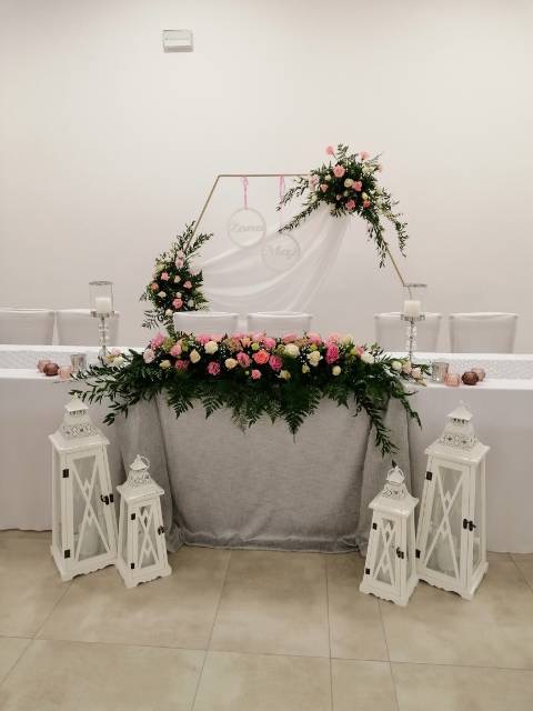 Dekoracja stołu weselnego z kwiatami różowych i białych róż, zielenią, białymi lampionami i geometrycznym złotym stelażem z imionami na białym tle.