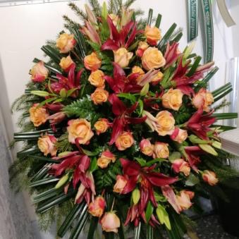 Duża kompozycja kwiatowa z pomarańczowych róż i ciemnoczerwonych lilii, ułożona na ciemnozielonych gałązkach, widoczna wstążka z zielonym napisem w tle po prawej stronie.
