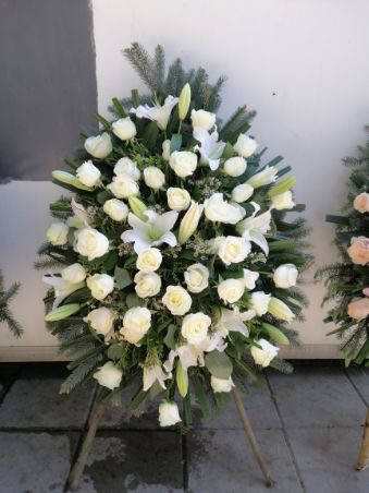 Kompozycja kwiatowa z białych róż i lilii na drewnianym stojaku, z elementami świerku, na tle szarej ściany i kostki brukowej.