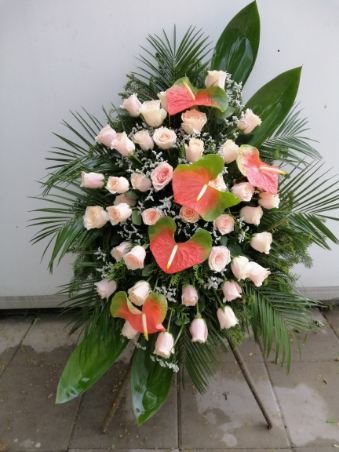 Kompozycja kwiatowa na stojaku: bladoróżowe róże, anturium z zielonymi liśćmi i palmowe liście na jasnym tle.