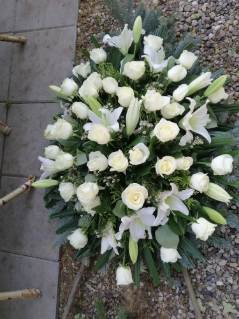 Kompaktowa kompozycja kwiatowa z białych róż i lilii, ułożona na ciemnozielonym podkładzie z gałązek, widoczna z góry na tle szarej kostki brukowej.