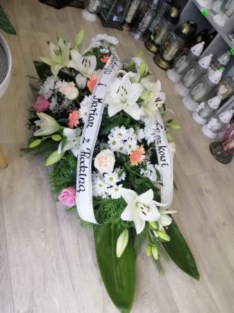 Kompozycja kwiatowa z białych lilii, róż i margaretek, przewiązana białą wstęgą z czarnym napisem żałobnym, ułożona na podłożu z zielonych liści.