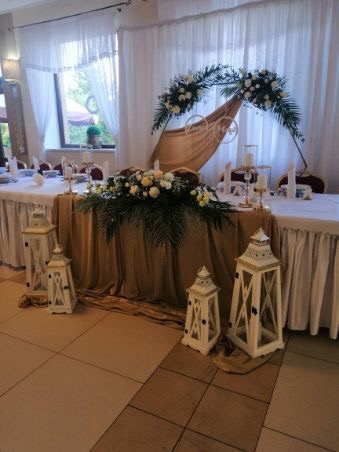 Udekorowany stół weselny z białymi i kremowymi kwiatami, zielonymi liśćmi, złotą tkaniną i białymi lampionami, ustawiony w sali z białymi zasłonami.