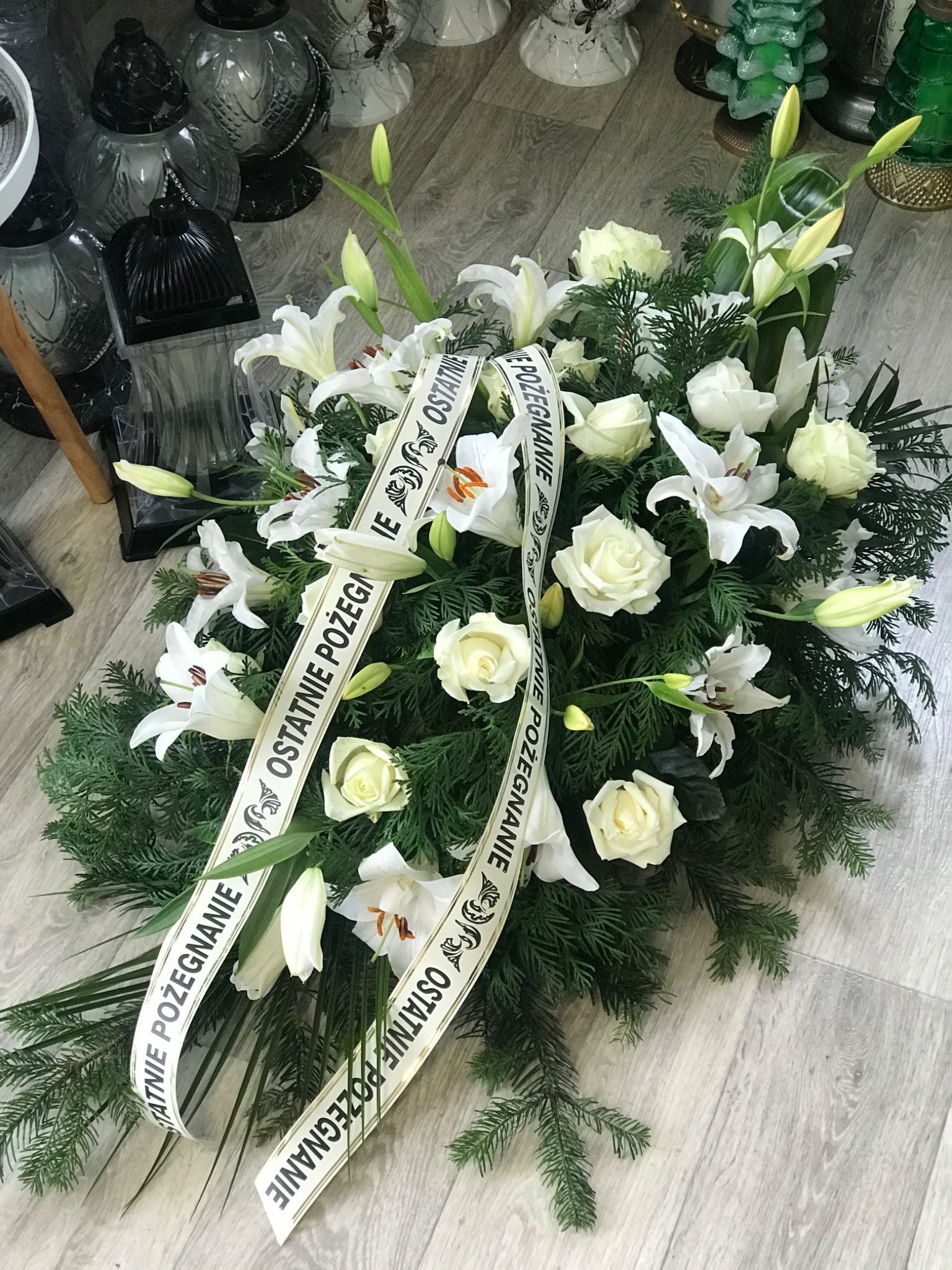 Kompozycja kwiatowa z białych róż i lilii, przewiązana kremową wstążką z napisem 'Ostatnie Pożegnanie', leżąca na podłodze z widocznymi lampionami w tle.