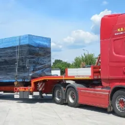 Czerwony ciągnik siodłowy Scania z naczepą niskopodwoziową przewożący zabezpieczony ładunek w postaci dużych, czarnych bloków owiniętych niebieską folią, na tle pochmurnego nieba.