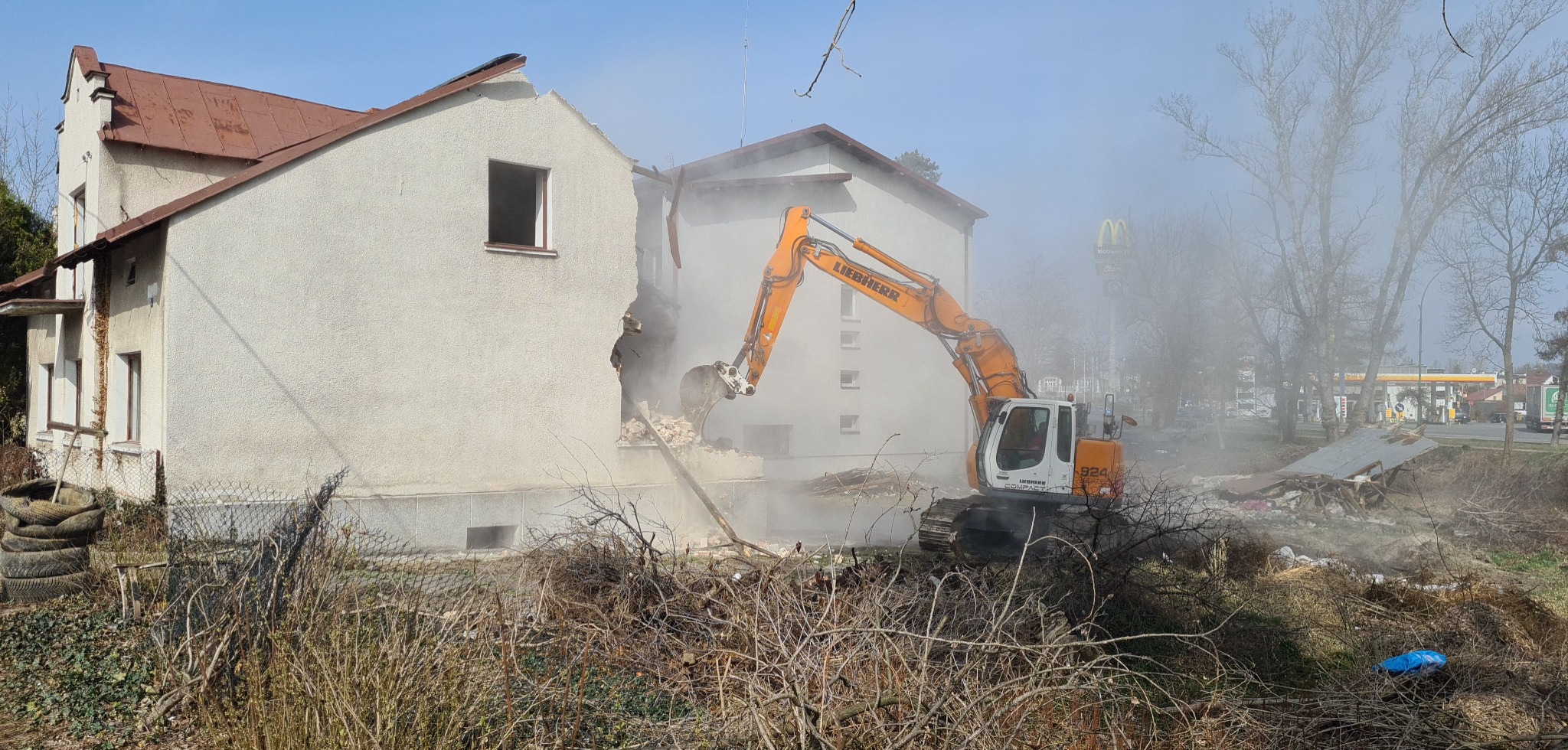 Pomarańczowo-biała koparka Liebherr 924 Compact burzy fragment ściany budynku z widocznymi cegłami, w tle budynki, drzewa i logo McDonald's, unoszący się pył.