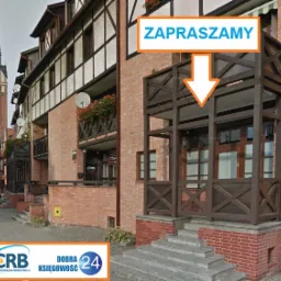 Fasada biura rachunkowego w Elblągu z drewnianym balkonem i napisem 'Zapraszamy' oraz logo firmy CRB Dobra Księgowość 24 na pierwszym planie, w tle fragment starówki z wysoką wieżą.