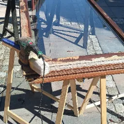 Renowacja ramy okiennej: szlifierka usuwa starą farbę z drewnianej ramy okna, stojącej na kozłach. Widoczne odpryski farby i surowe drewno.