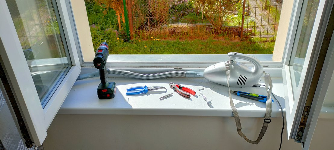 Otworzone okno z narzędziami: wiertarka, odkurzacz, kombinerki, nożyk. Widok na zewnątrz, na zieleń. Przygotowanie do naprawy lub konserwacji okna.