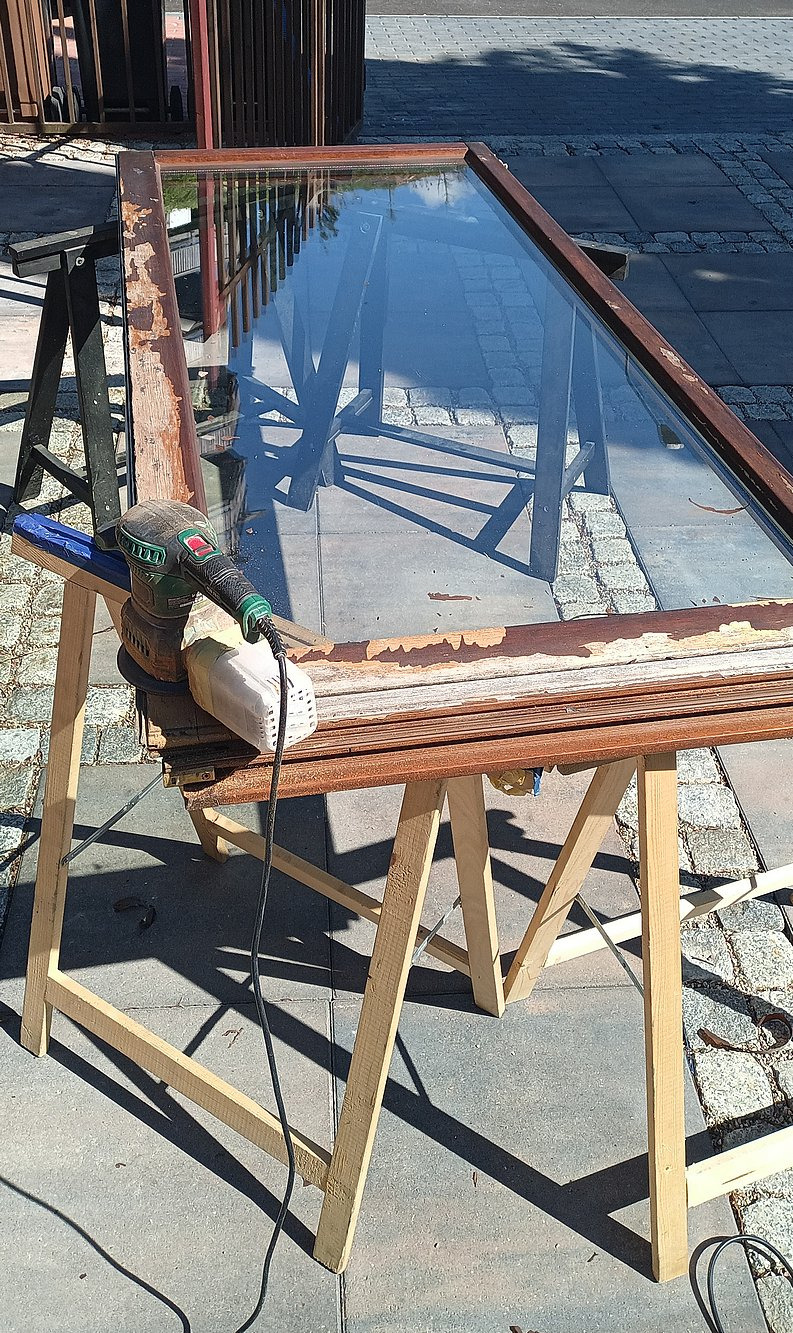 Renowacja ramy okiennej: szlifierka usuwa starą farbę z drewnianej ramy okna, stojącej na kozłach. Widoczne odpryski farby i surowe drewno.