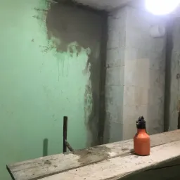 Fragment ściany z nałożoną warstwą szarej zaprawy, widoczne zacieki, na rusztowaniu stoi pomarańczowa butelka z rozpylaczem, oświetlenie górne.