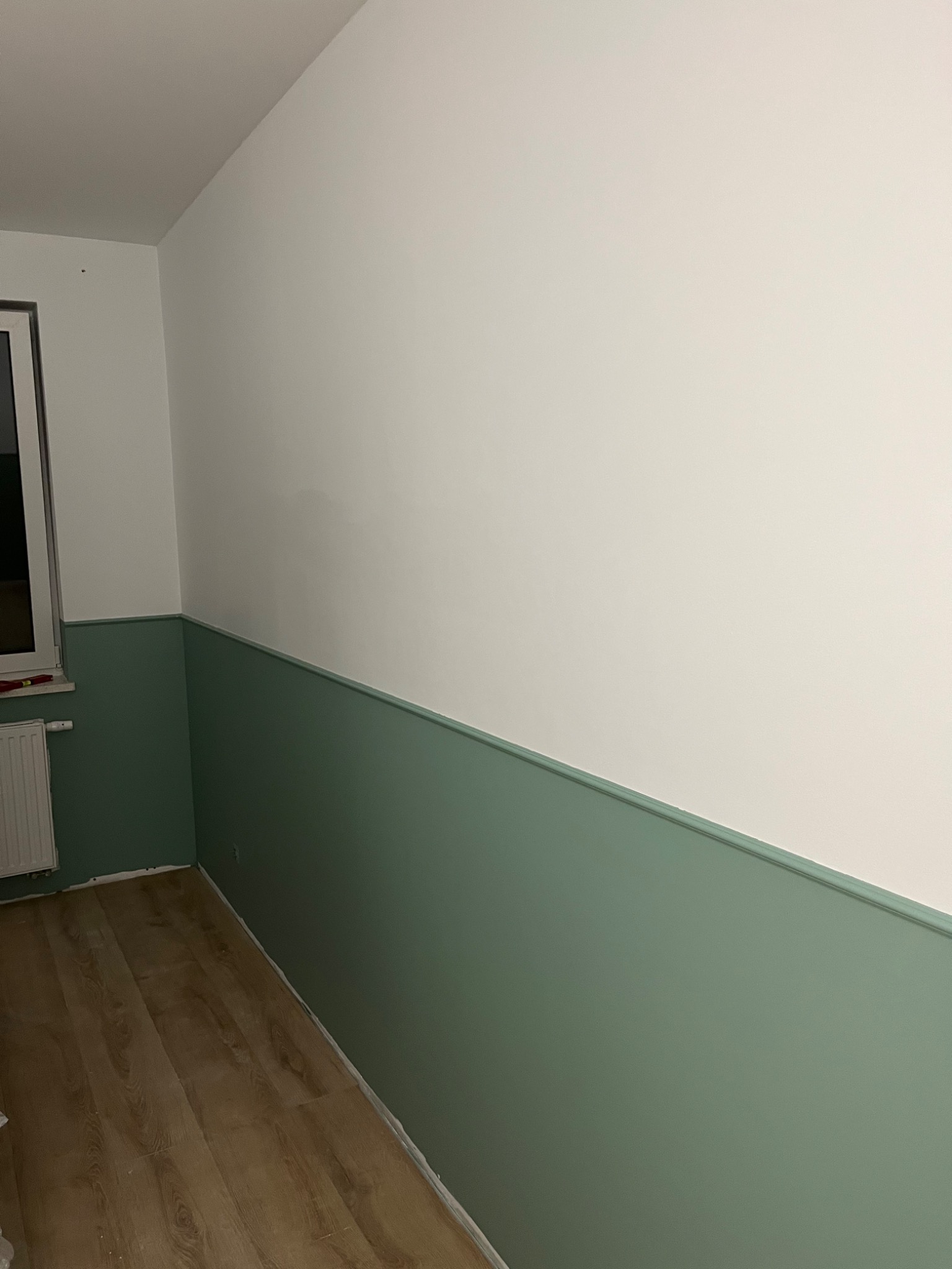 Pokój po remoncie z dwukolorowymi ścianami: górna część biała, dolna pomalowana na odcień zieleni, widoczna drewniana podłoga i grzejnik.