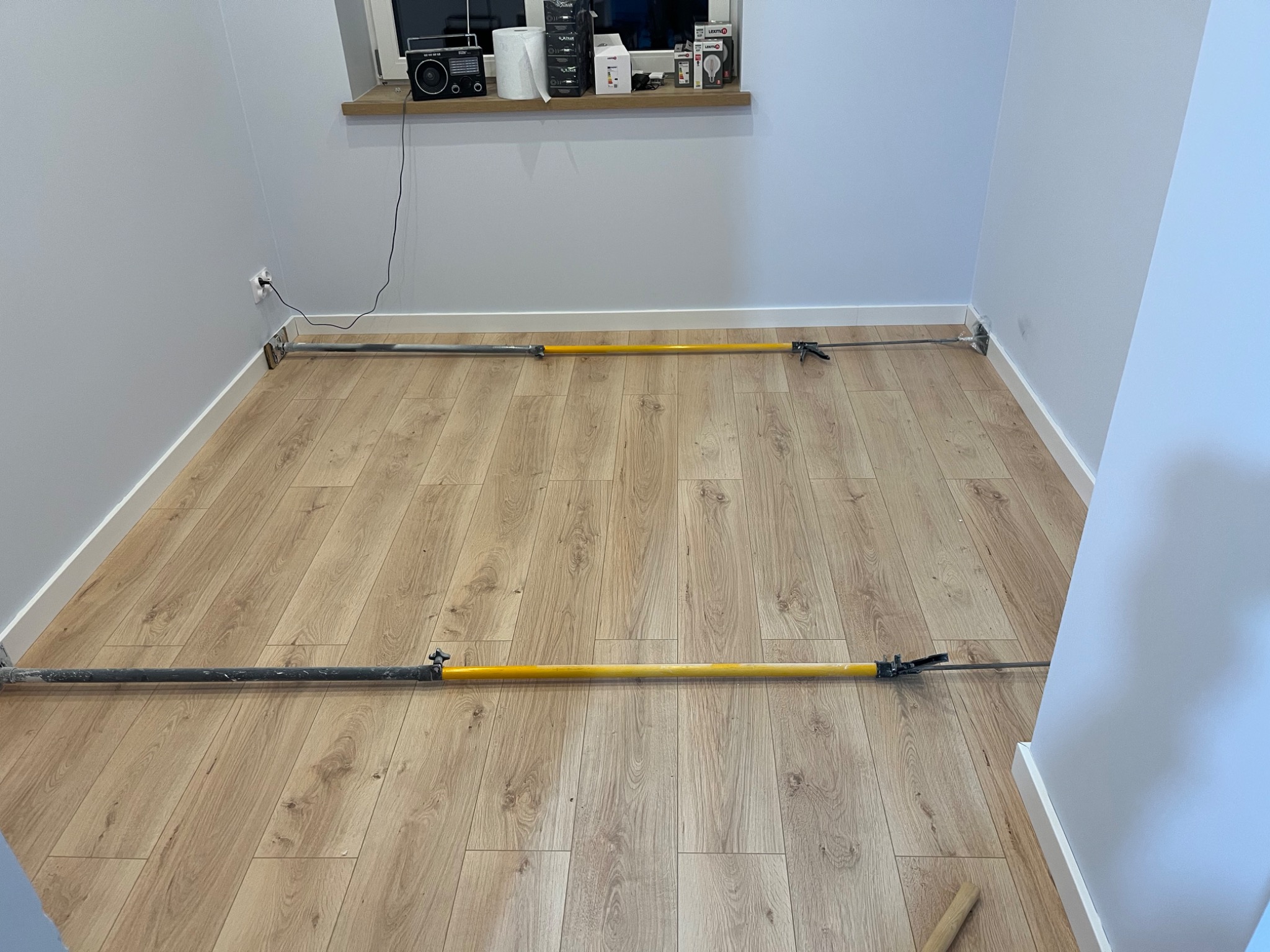 Pomieszczenie z panelami podłogowymi w trakcie montażu listew przypodłogowych przy użyciu specjalistycznego narzędzia zaciskowego z długim uchwytem.