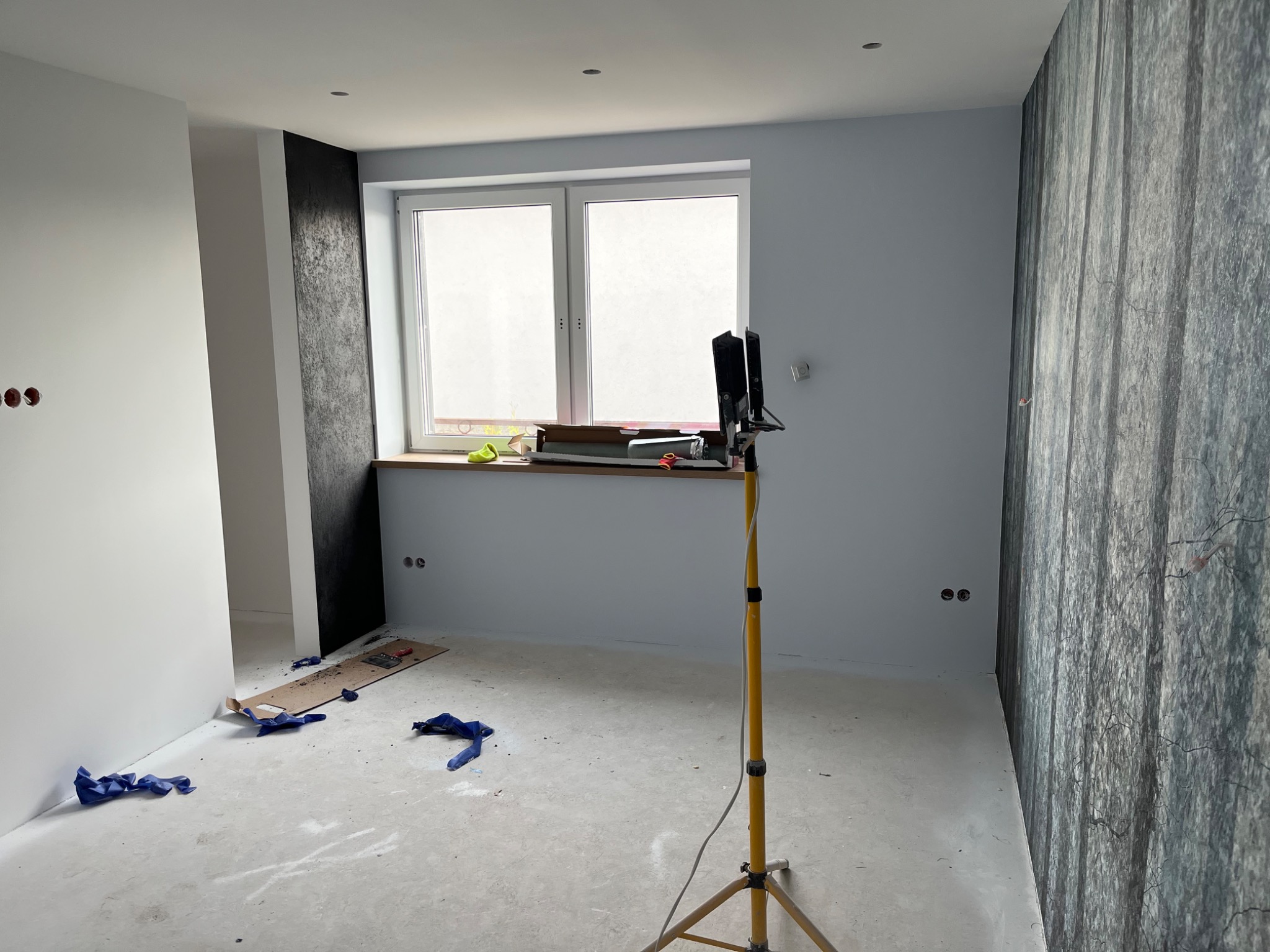 Pokój w trakcie prac wykończeniowych: ściany częściowo pomalowane na szaro, jedna ściana z tapetą w leśny motyw, żółty statyw oświetleniowy, resztki kartonu i niebieskich rękawic na podłodze, okno...