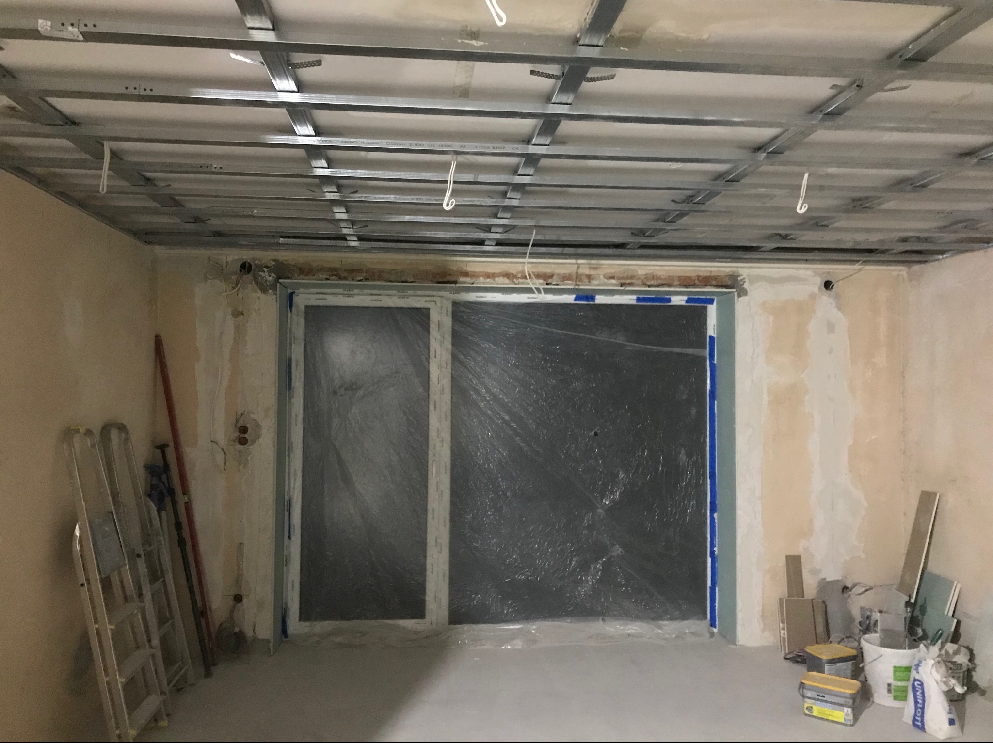 Wnętrze w trakcie prac: metalowa konstrukcja podwieszanego sufitu, zabezpieczone folią okno, drabina oparta o ścianę, widoczne materiały budowlane.