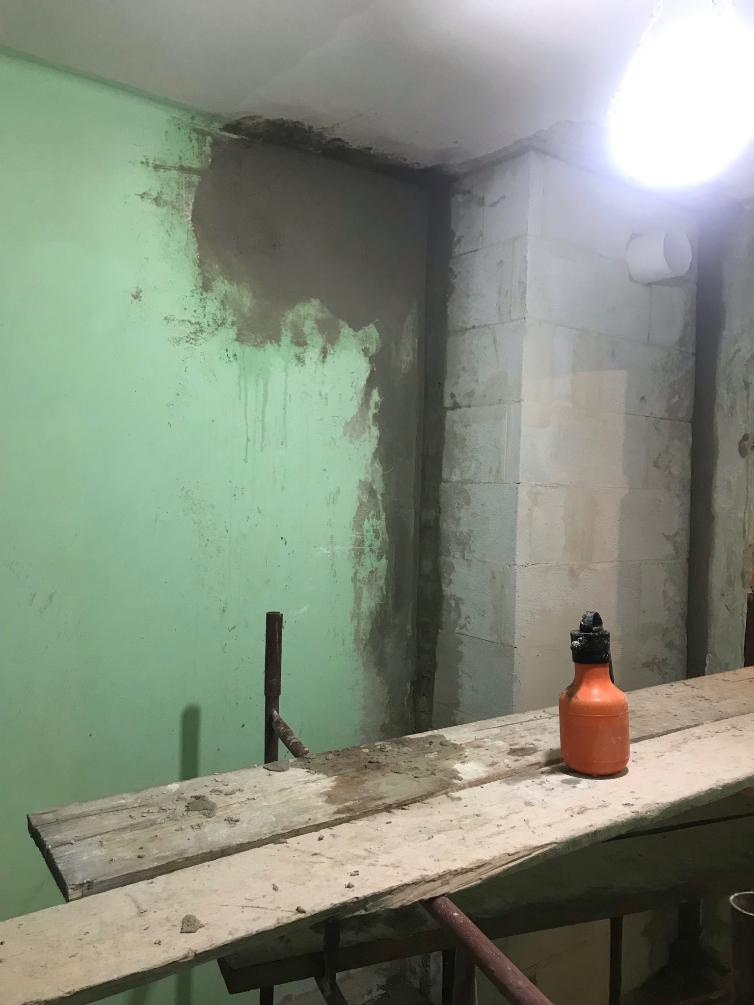 Fragment ściany z nałożoną warstwą szarej zaprawy, widoczne zacieki, na rusztowaniu stoi pomarańczowa butelka z rozpylaczem, oświetlenie górne.