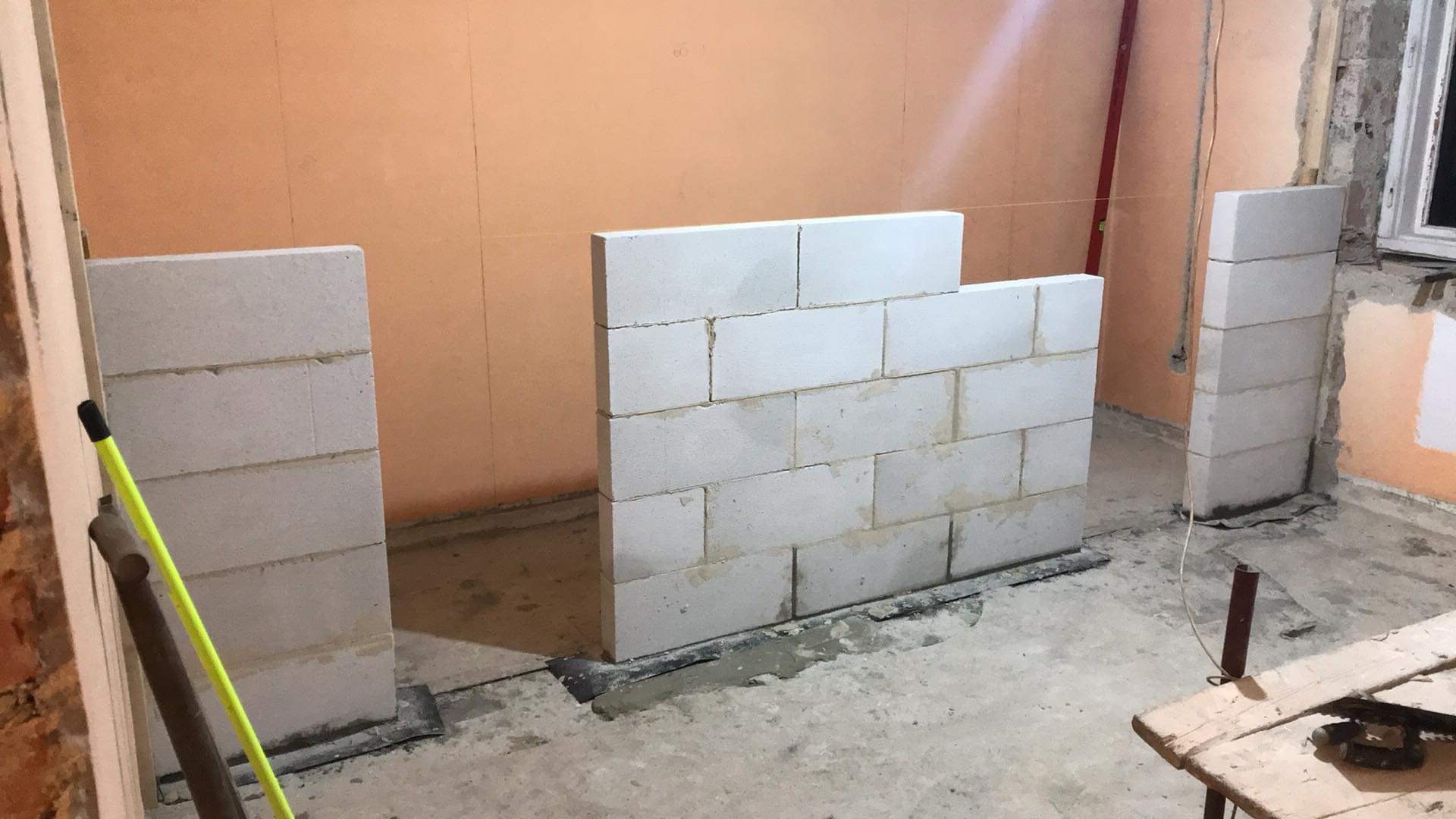 Wznoszenie ściany działowej z bloczków betonu komórkowego w trakcie prac wykończeniowych w pomieszczeniu z pomarańczowymi ścianami. Widoczny sznurek wyznaczający linię ściany, zaprawa oraz narzędzia.