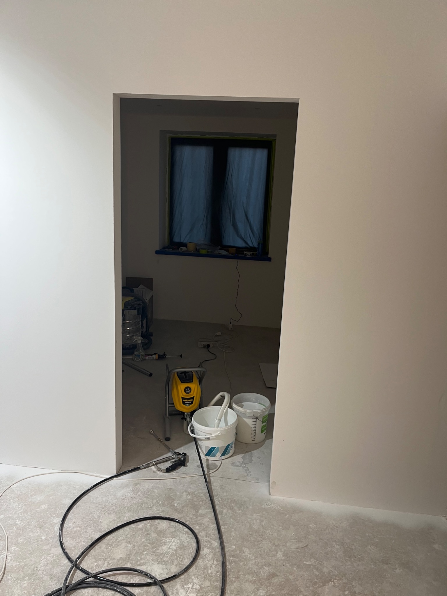 Pokój w trakcie malowania natryskowego, widoczny agregat malarski Wagner, dwa wiadra z farbą, okno zasłonięte folią malarską, kabel agregatu zwinięty na podłodze.