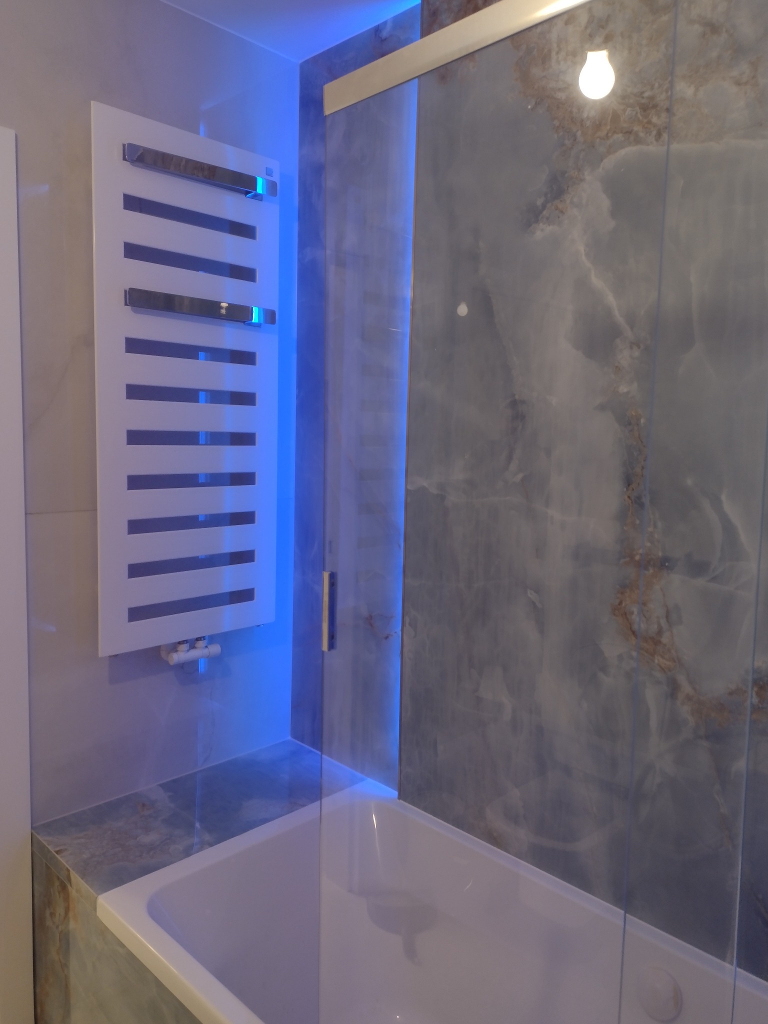 Nowoczesna łazienka z wanną obłożoną marmurową płytą, grzejnikiem drabinkowym i podświetleniem LED.