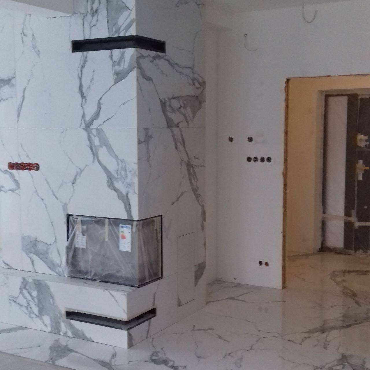 Wykończony kominek obłożony płytami imitującymi marmur, z czarnym wkładem kominkowym zabezpieczonym folią, widoczne gniazdka elektryczne i otwór drzwiowy w tle.