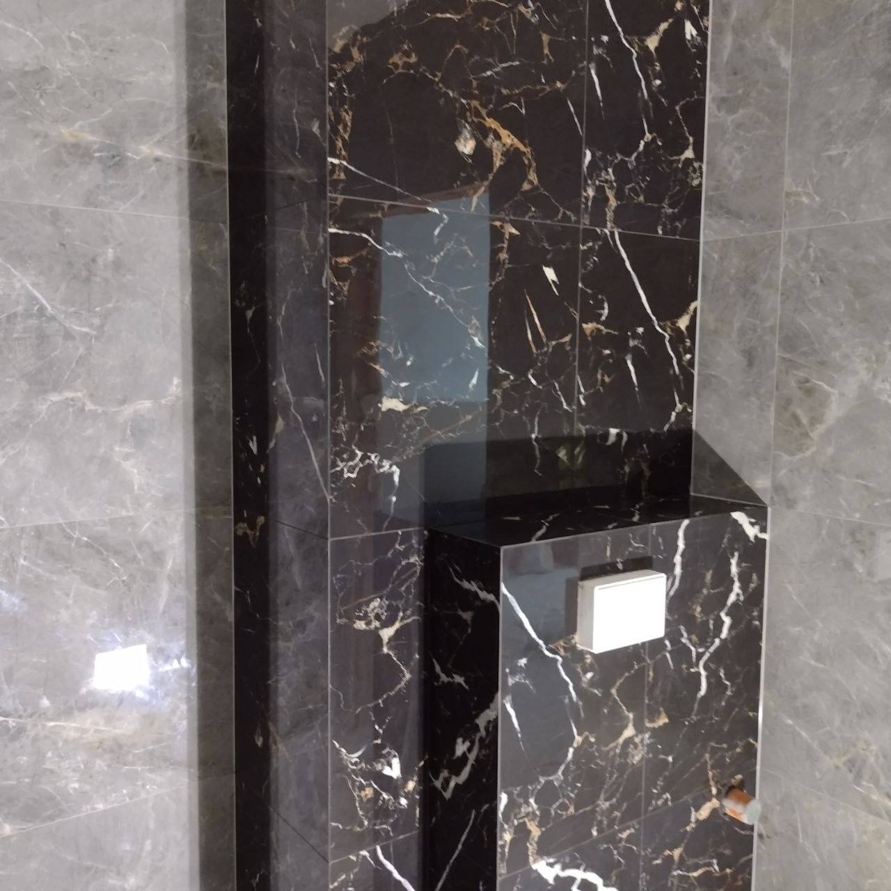 Wykończona wnęka w łazience z czarnych płytek imitujących marmur z białymi i złotymi żyłkami, sąsiadująca z szarymi płytkami, widoczny biały przycisk spłuczki.