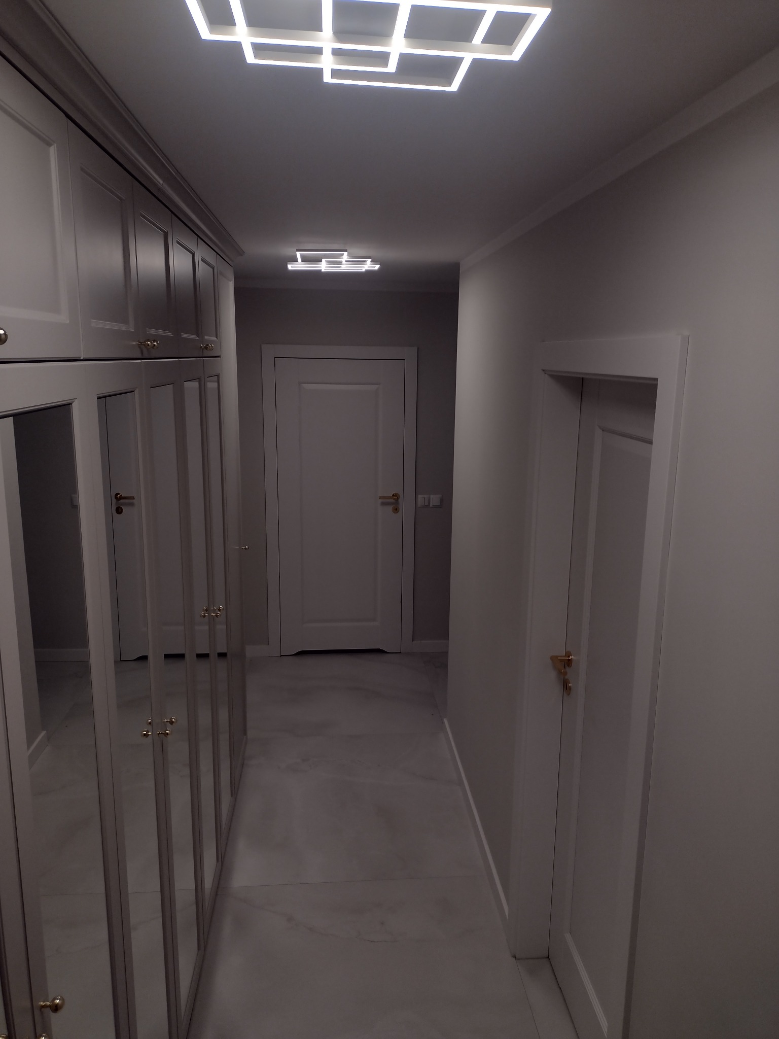 Nowoczesny korytarz w odcieniach szarości z szafą w zabudowie z lustrzanymi frontami, oświetleniem sufitowym LED w geometryczne wzory i marmurową posadzką.
