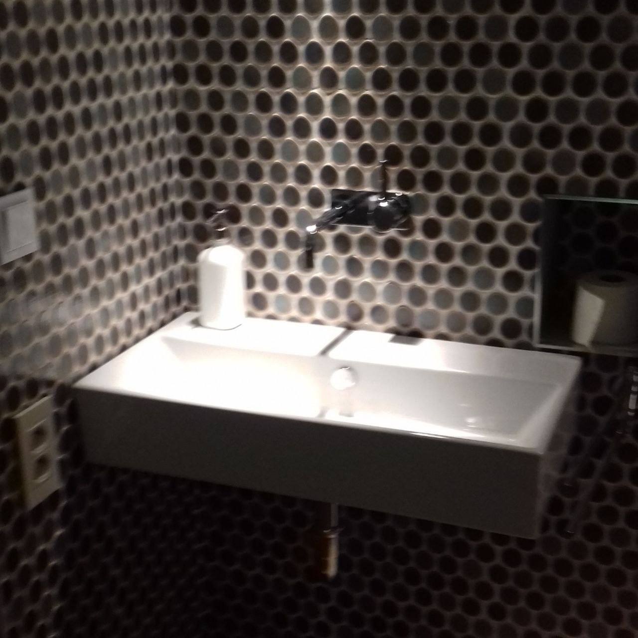 Nowoczesna łazienka z umywalką nablatową i okrągłymi, mozaikowymi płytkami w odcieniach szarości i czerni, chromowana bateria, dozownik mydła i półka z papierem toaletowym.