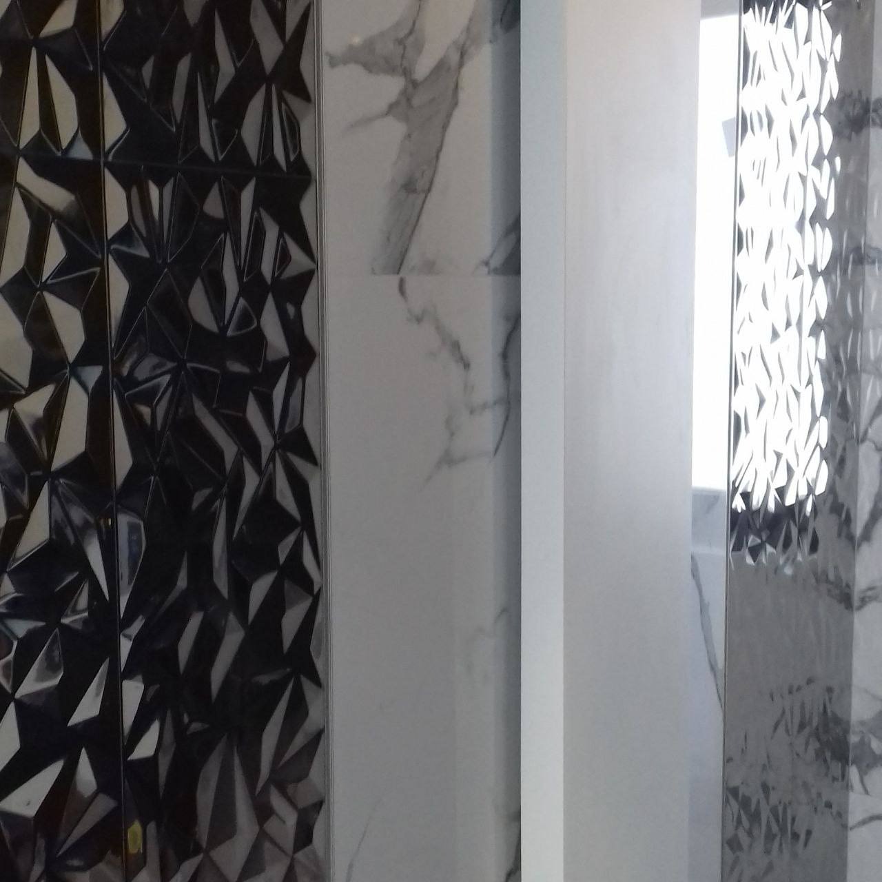 Fragment ściany z różnorodnymi płytkami: czarne lustrzane z geometrycznym wzorem, białe marmurowe oraz białe gładkie, oddzielone pionowymi pasami.