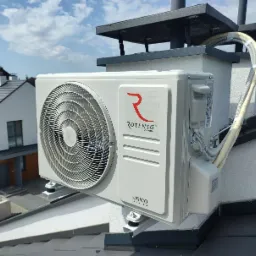 Zewnętrzna jednostka klimatyzacji Rotenso Unico Series zamontowana na dachu budynku mieszkalnego, widoczne przewody i komin wentylacyjny, w tle zabudowa jednorodzinna i błękitne niebo.