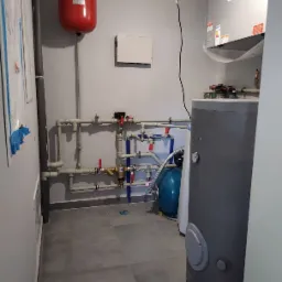 Wnętrze kotłowni z szarym bojlerem, czerwonym zbiornikiem wyrównawczym, niebieskim zbiornikiem hydroforowym oraz systemem rur z zaworami.