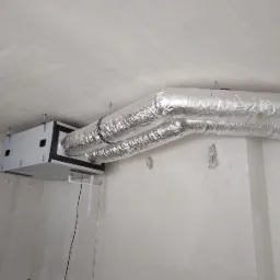 Centrala wentylacyjna z dwoma przewodami wentylacyjnymi w srebrnej izolacji, zamontowana pod sufitem w surowym pomieszczeniu.