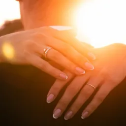 Dłonie kobiety obejmujące mężczyznę, widoczne pierścionki zaręczynowy i obrączka, ciepłe światło zachodzącego słońca.