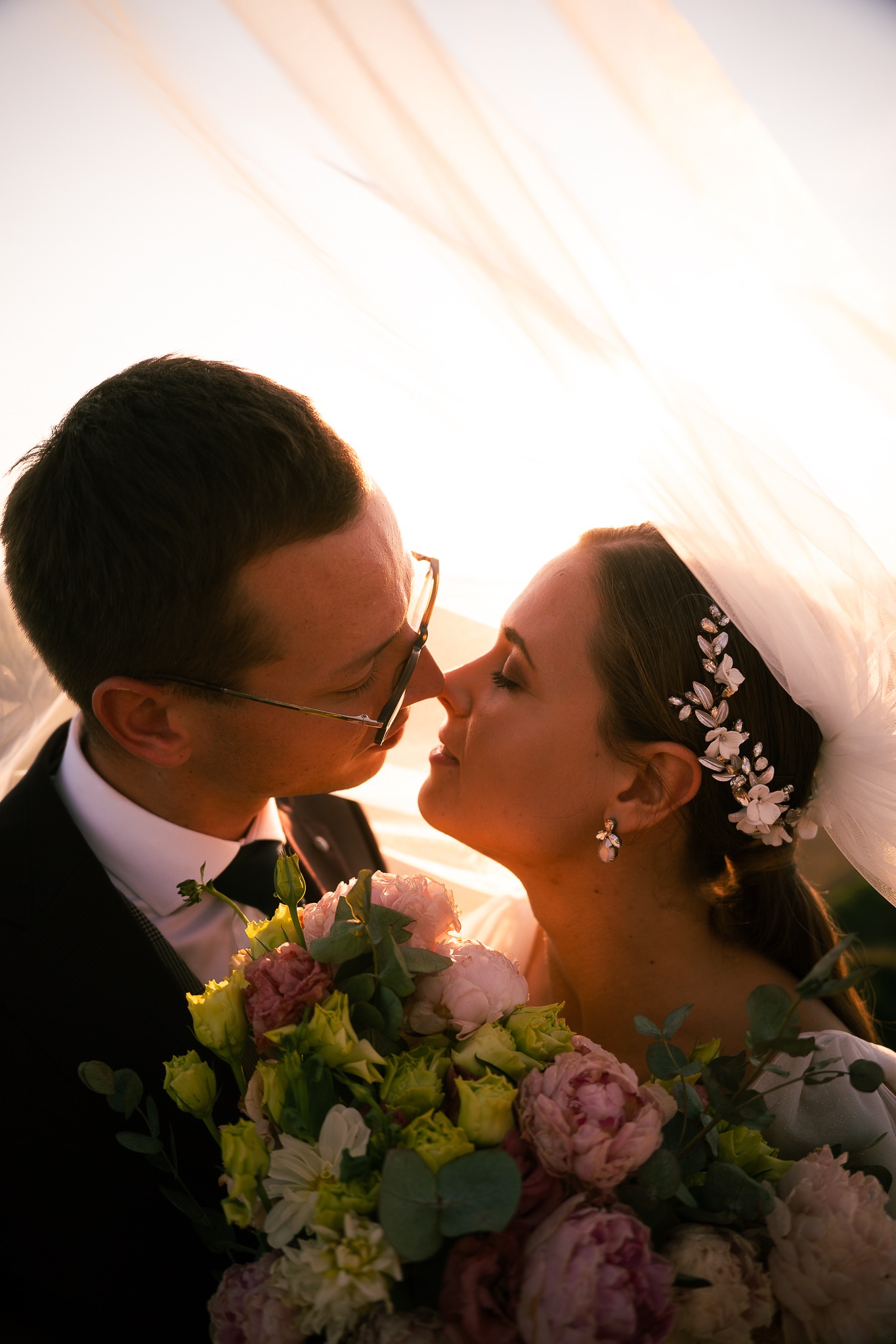 Para młoda tuż przed pocałunkiem, on w okularach, ona z ozdobą we włosach, oboje pod welonem, trzymają bukiet kwiatów w odcieniach różu i zieleni, ciepłe światło zachodzącego słońca.