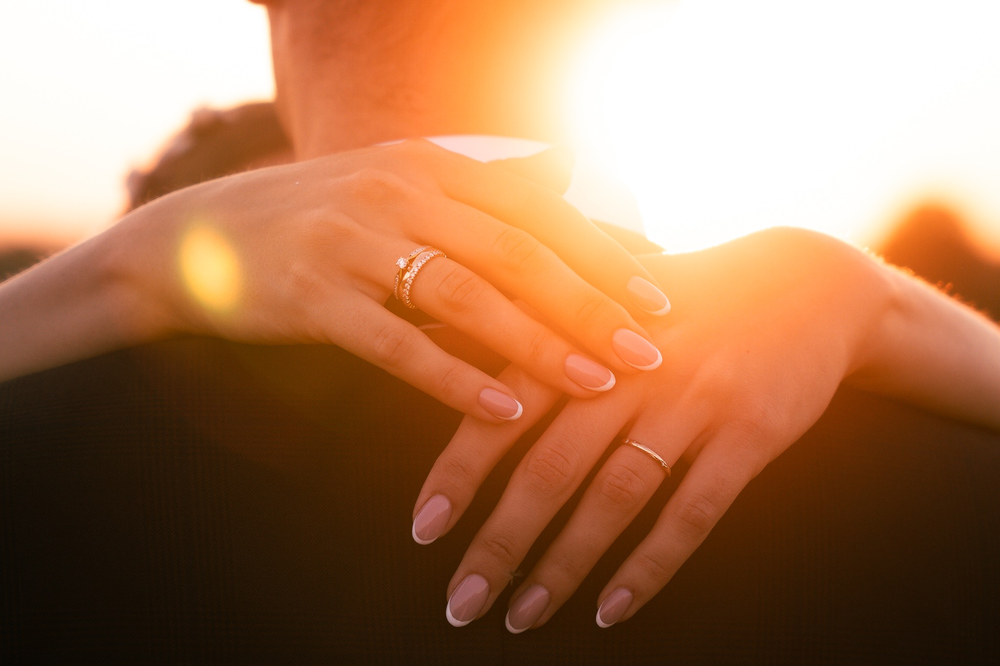 Dłonie kobiety obejmujące mężczyznę, widoczne pierścionki zaręczynowy i obrączka, ciepłe światło zachodzącego słońca.