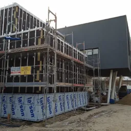 Budynek w trakcie ocieplania i montażu elewacji, widoczne rusztowania, płyty izolacyjne i okładzinowe Larson, stos materiałów budowlanych obok budynku.